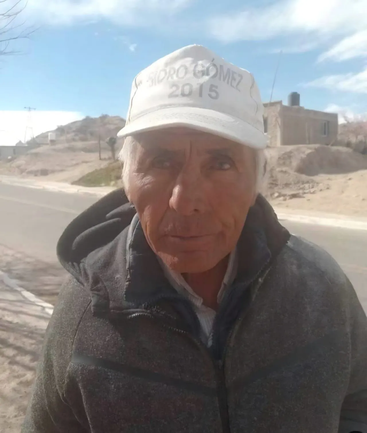 Un hombre mayor con gorra blanca y campera gris parado al costado de un camino de tierra.