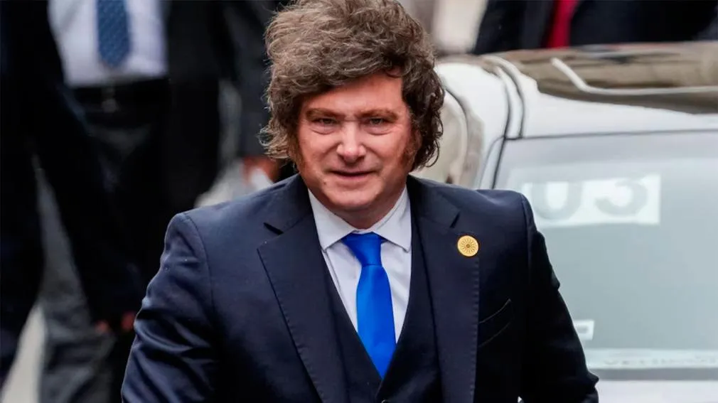 Javier Milei viste traje oscuro con chaleco, corbata azul y pin dorado en la solapa mientras camina.